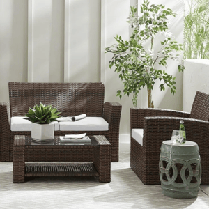 Outdoor furniture set featuring a wicker loveseat, two chairs with white cushions, and a matching coffee table, complemented by potted plants and a small decorative side table.