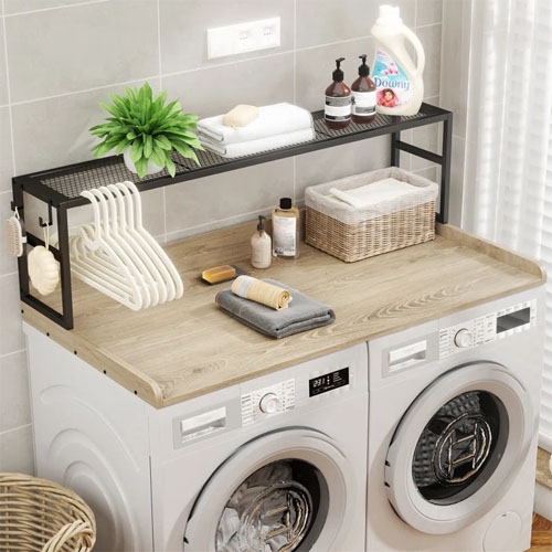 A compact laundry setup featuring a wooden countertop over a washer and dryer, with a black metal shelf holding detergent, baskets, cleaning supplies, hangers, and a decorative plant.
