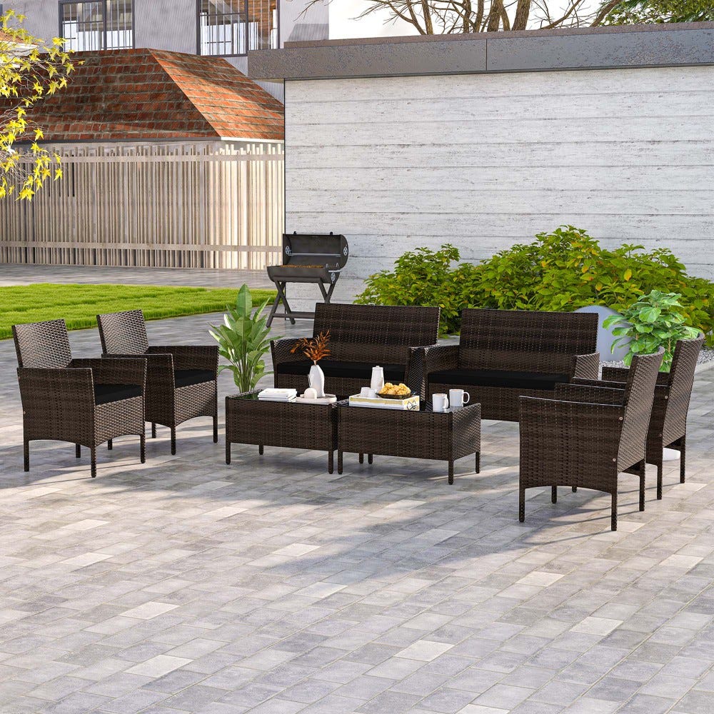 The 8pc Costway Patio Set in black features wicker furniture, including four armchairs, two loveseats, and two coffee tables, arranged on a patio. The set includes cushions and two decorative plants, with a BBQ grill visible in the background.