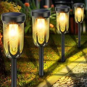 Four solar-powered garden lights illuminate a pathway, with warm yellow light encased in cylindrical glass housing mounted on black stakes along a landscaped garden area.