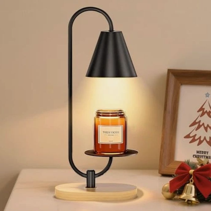 A modern candle warmer lamp with a black metal frame and wooden base is illuminating a candle. A framed Christmas-themed picture with decorations is placed nearby.
