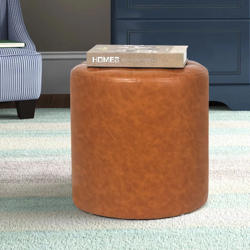 A cylindrical, tan faux-leather ottoman with a smooth texture, placed on a striped carpet, and topped with a book.