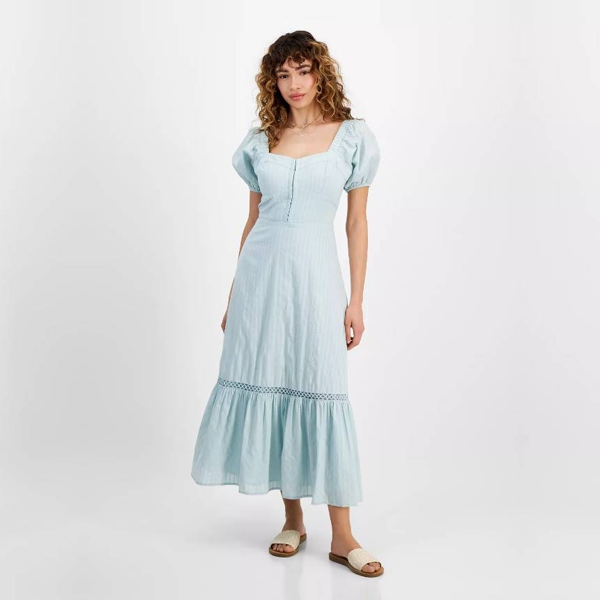 A woman wearing a light blue midi dress with puff sleeves and decorative trim, paired with simple sandals.