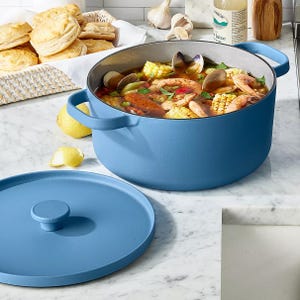 A blue Dutch oven filled with a seafood boil, including shrimp, clams, corn, and herbs, is shown on a marble countertop next to a lid, with biscuits and kitchen items in the background.