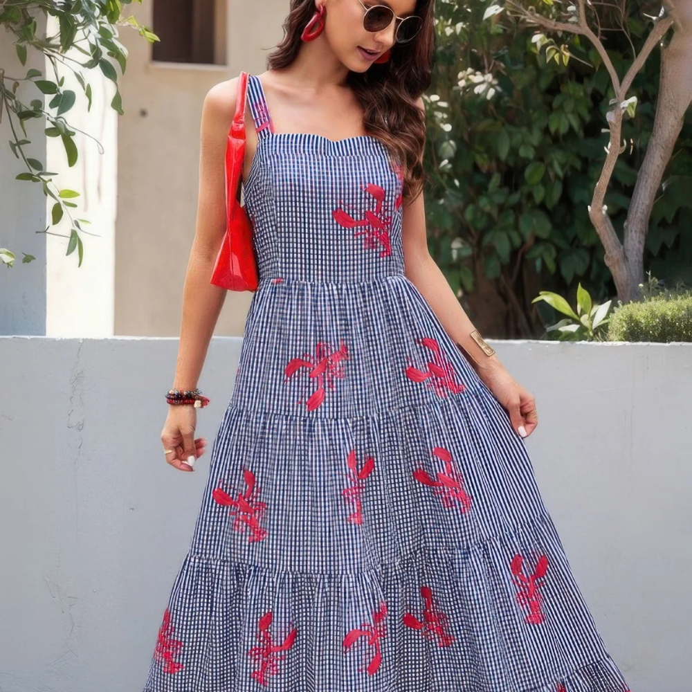 A woman in a blue and white gingham dress with red patterns, accessorized with round sunglasses, red earrings, and a red bag. She's wearing bracelets and standing near a wall with greenery in the background.