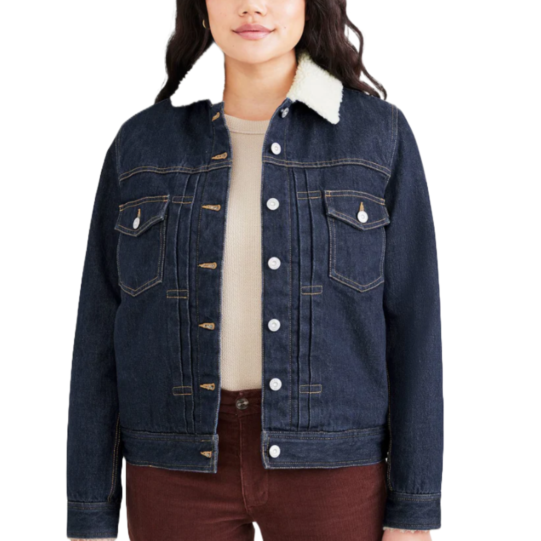 Dark blue denim trucker jacket with a white sherpa collar, featuring button closures and two chest pockets with flap buttons. Worn over a beige top and paired with brown pants.