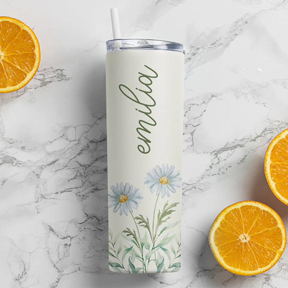A personalized tumbler with a straw, featuring the name \“emilia\“ and a floral design with daisies, placed on a marble surface beside halved oranges.