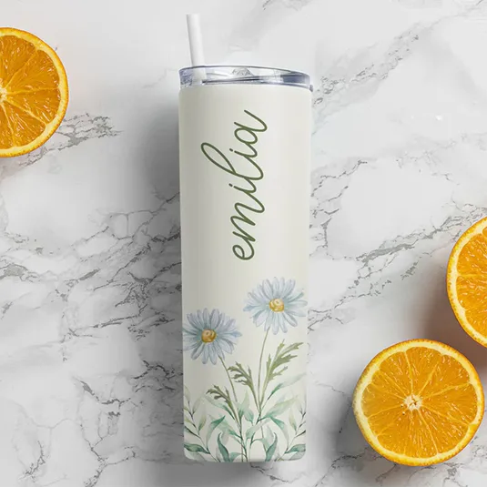 A personalized tumbler with a straw, featuring the name \“emilia\“ and a floral design with daisies, placed on a marble surface beside halved oranges.