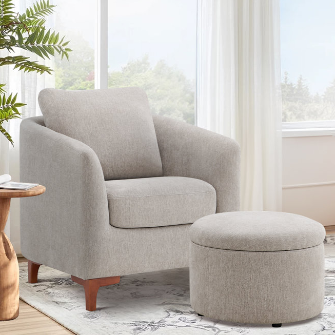 A light gray armchair with a matching round ottoman is situated on a patterned rug beside a wooden side table, with a leafy plant and window in the background.