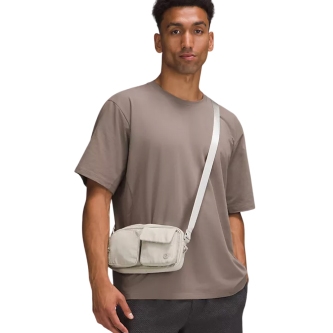 A person wears a taupe t-shirt and a light-colored crossbody bag with two front pockets, paired with dark pants.