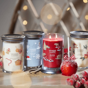 Four Yankee Candle jars with holiday-themed designs: Munich Christmas Market, North Pole Highway, Big Apple Christmas, and London Christmas Star. A red candy apple with a ribbon is placed in front.
