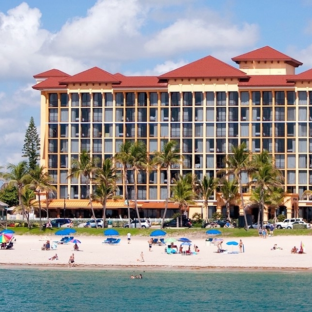 Beachfront resort building with palm trees and people on the beach under umbrellas.