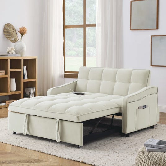 A light-colored, tufted convertible sofa bed with nailhead accents, shown in a living room setting, featuring a side pocket for storage and a wooden bookshelf nearby.