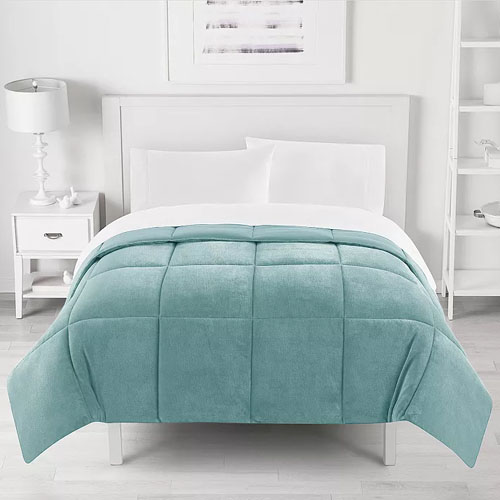 A neatly made bed features a light teal checkered comforter with white sheets, a white headboard, a white nightstand with a lamp on the left, and shelves on the wall to the right.
