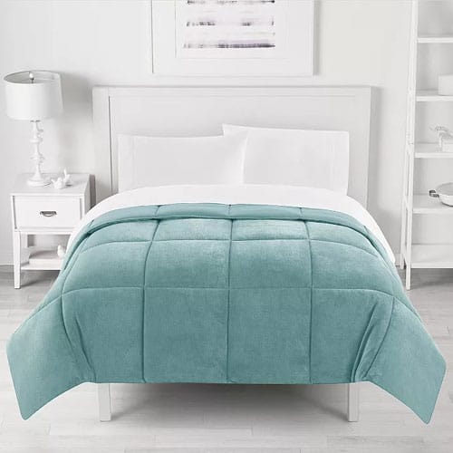 A neatly made bed features a light teal checkered comforter with white sheets, a white headboard, a white nightstand with a lamp on the left, and shelves on the wall to the right.
