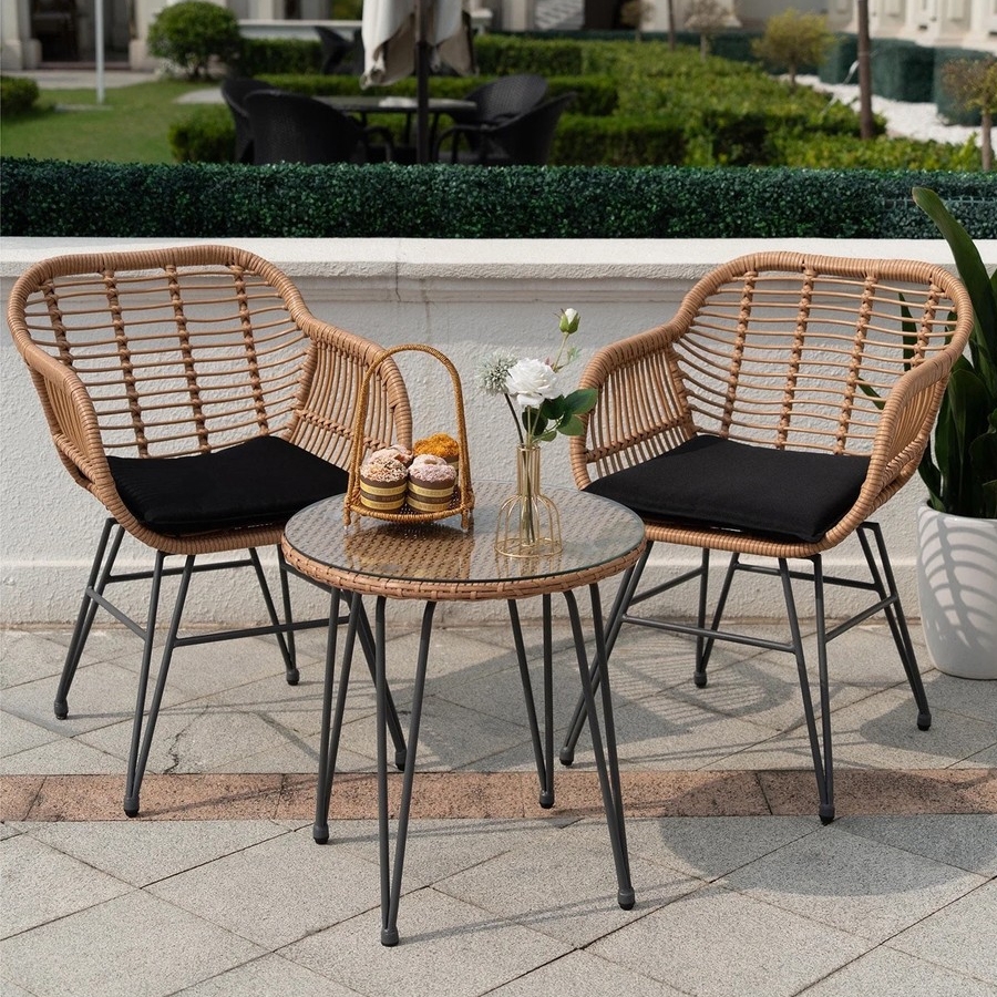 A 3-piece patio bistro set featuring two rattan chairs with black cushions and a round glass-top rattan table. The setup includes a small basket with macarons and a vase with white flowers.