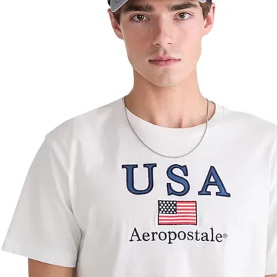 A person is wearing a white Aeropostale t-shirt with \“USA\“ and an American flag, along with a silver necklace and a gray cap.