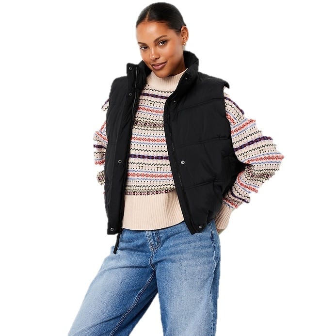 A woman is wearing a black puffer vest over a multicolored patterned sweater paired with blue jeans.