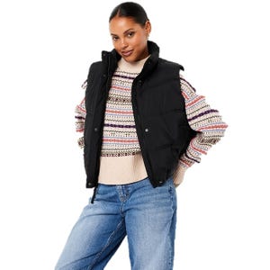 A woman is wearing a black puffer vest over a multicolored patterned sweater paired with blue jeans.