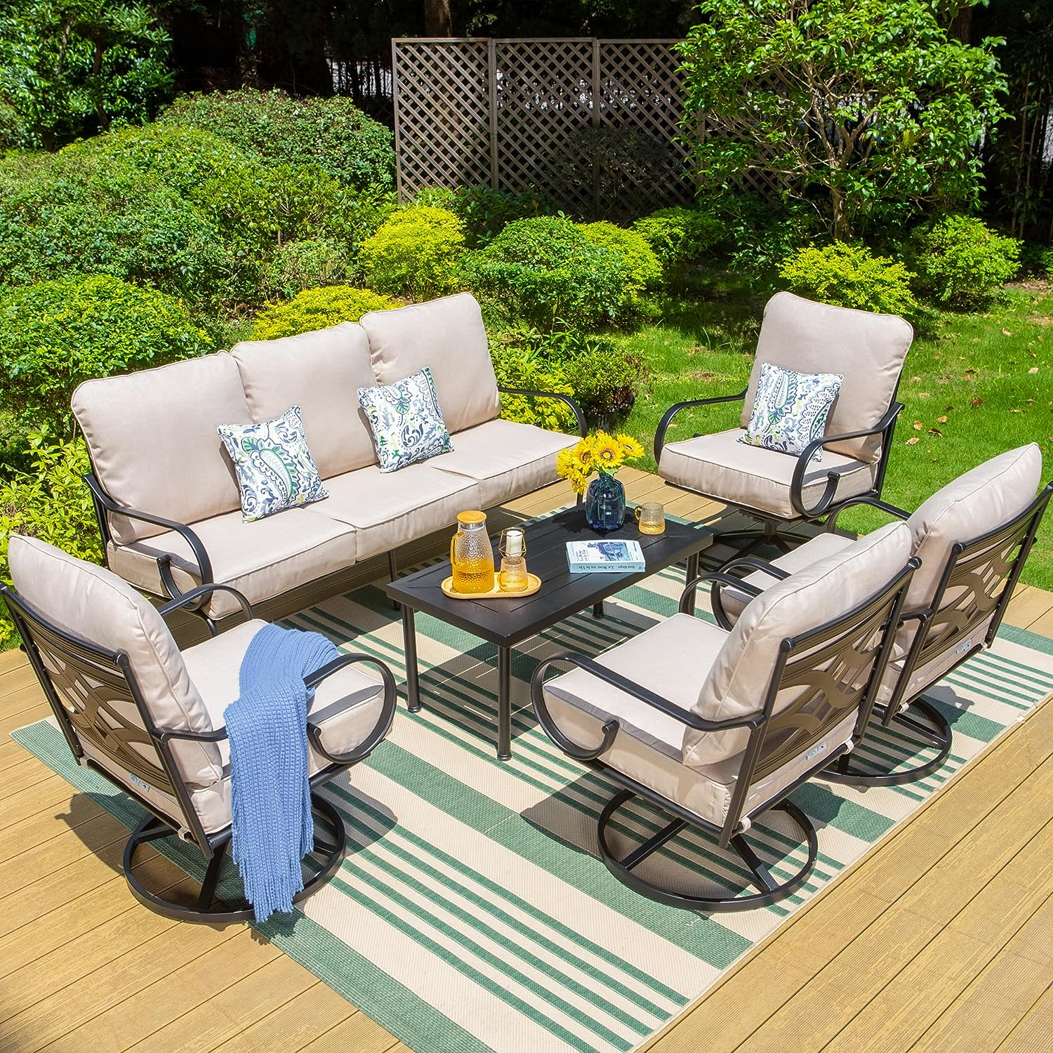 Patio furniture set with beige cushions, including a three-seater sofa, four swivel chairs, a coffee table on a striped outdoor rug, and decorative pillows, set in a garden area.