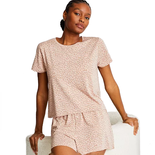 A person is wearing a pastel pink leopard print pajama set consisting of a short-sleeve top and matching shorts, leaning against a backrest.