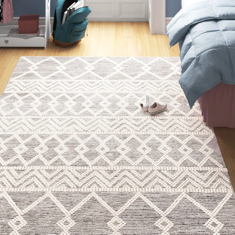 A geometric-patterned area rug with neutral tones is placed on a light wood floor near a bed with a blue comforter. A pair of casual slippers is shown on the rug.