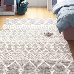 A geometric-patterned area rug with neutral tones is placed on a light wood floor near a bed with a blue comforter. A pair of casual slippers is shown on the rug.