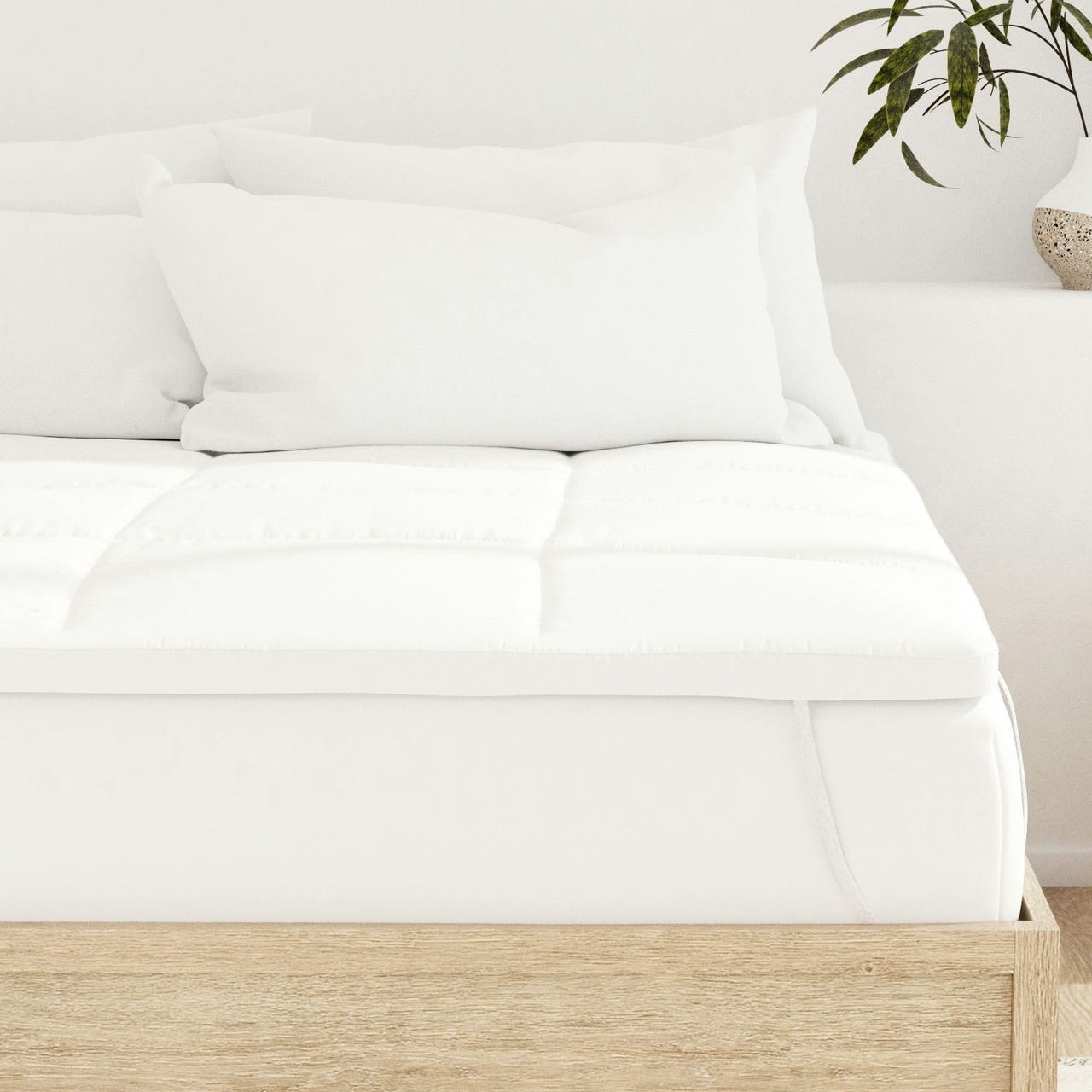A bed with a white mattress topper and several white pillows on a light wooden frame is shown. A potted plant is visible on a side surface.