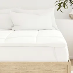 A bed with a white mattress topper and several white pillows on a light wooden frame is shown. A potted plant is visible on a side surface.