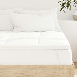 A bed with a white mattress topper and several white pillows on a light wooden frame is shown. A potted plant is visible on a side surface.