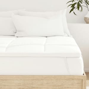 A bed with a white mattress topper and several white pillows on a light wooden frame is shown. A potted plant is visible on a side surface.