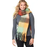 A woman wearing a ribbed gray sweater paired with a large, colorful plaid scarf featuring shades of green, orange, and brown. The scarf has long fringe along the edges.