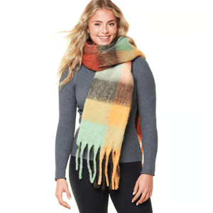 A woman wearing a ribbed gray sweater paired with a large, colorful plaid scarf featuring shades of green, orange, and brown. The scarf has long fringe along the edges.