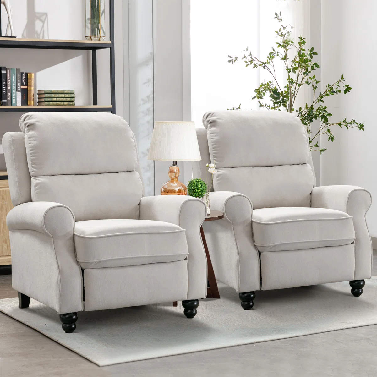 Two light gray upholstered recliners with rounded armrests and dark wooden legs are arranged side by side. A small table with a lamp and plant sits between them, and a bookshelf is visible in the background.