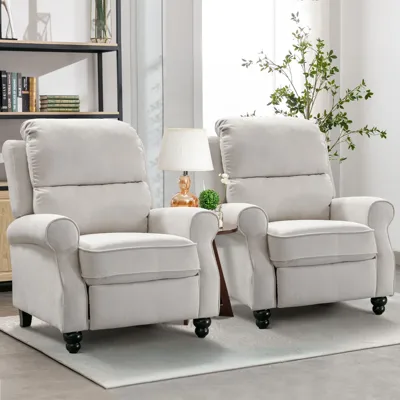 Two light gray upholstered recliners with rounded armrests and dark wooden legs are arranged side by side. A small table with a lamp and plant sits between them, and a bookshelf is visible in the background.