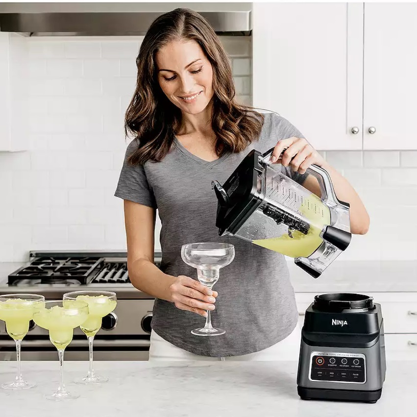 A woman is pouring a blended green beverage from a blender into a margarita glass, with multiple filled glasses on the counter and a Ninja blender base beside her.