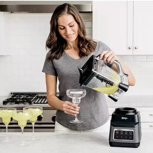 A woman is pouring a blended green beverage from a blender into a margarita glass, with multiple filled glasses on the counter and a Ninja blender base beside her.