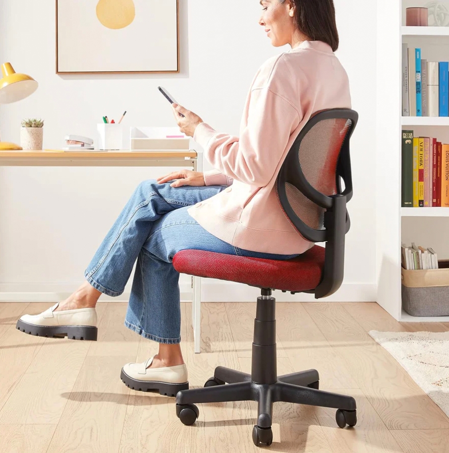 A woman is sitting on a red and black office chair with a mesh backrest, using a smartphone.