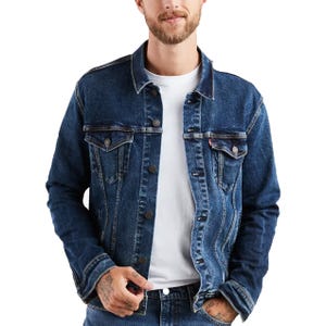 A man wears a classic Levi's denim trucker jacket, featuring a button-up front, dual chest pockets with flaps, and side pockets. The jacket is dark blue and worn over a white T-shirt, complemented by matching blue jeans.