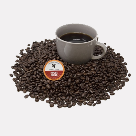 A pile of dark roasted coffee beans surrounding a cup of coffee next to a 96-count Pintail House Blend K-Cups package.