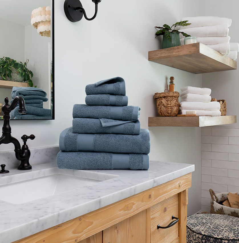 A stack of blue towels is neatly arranged on a marble bathroom counter, with additional towels and bath items on wooden shelves nearby.