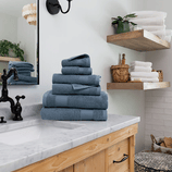 A stack of blue towels is neatly arranged on a marble bathroom counter, with additional towels and bath items on wooden shelves nearby.