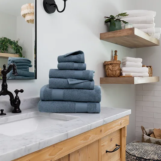 A stack of blue towels is neatly arranged on a marble bathroom counter, with additional towels and bath items on wooden shelves nearby.