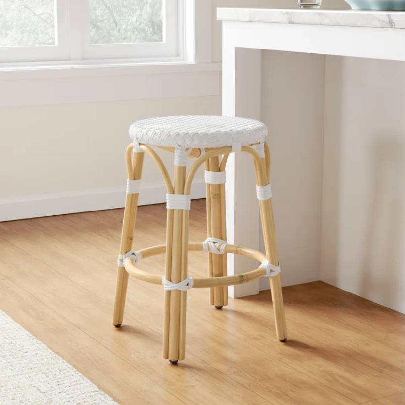 A natural wood counter stool with a round white woven seat and matching white bindings on the legs and footrest. It's positioned next to a white countertop with a light-colored wooden floor beneath.