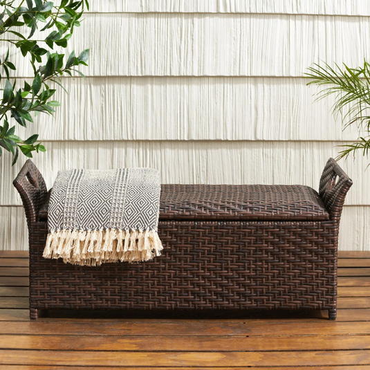 Wicker storage bench with built-in handles, featuring a gray geometric fringed throw on top. It is placed on a wooden floor with a white textured wall and plants in the background.