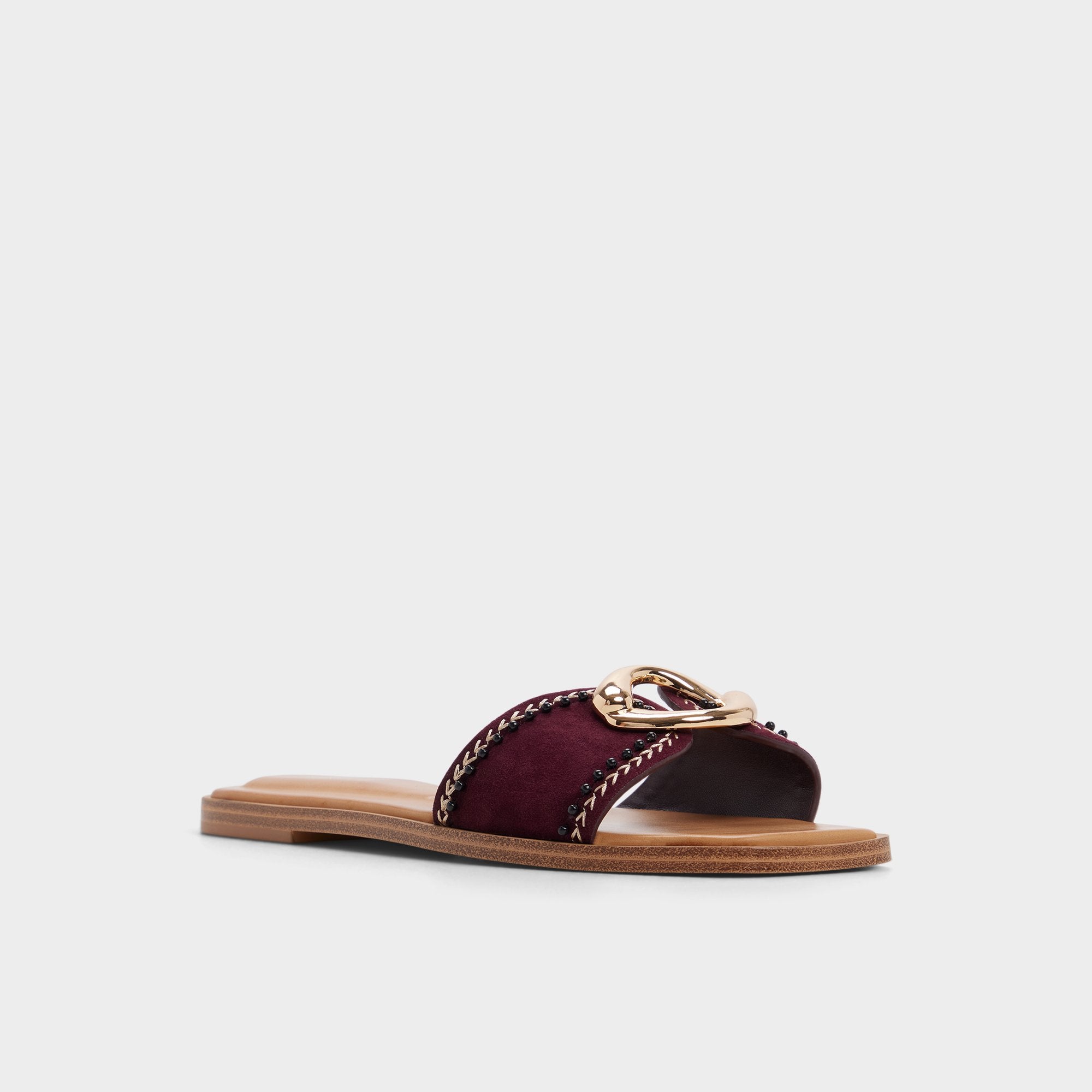 A burgundy slide sandal featuring a wide strap with chain trim and a gold metal accent, set on a flat, tan sole.