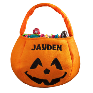 Orange Halloween-themed trick-or-treat basket with a smiling jack-o'-lantern face and the name \“Jayden\“ embroidered, filled with various candies.