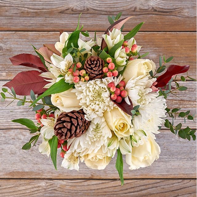 A bouquet featuring cream roses, white chrysanthemums, alstroemeria, pinecones, red hypericum berries, and greenery, arranged on a wooden surface for a rustic autumnal look.