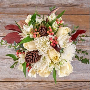 A bouquet featuring cream roses, white chrysanthemums, alstroemeria, pinecones, red hypericum berries, and greenery, arranged on a wooden surface for a rustic autumnal look.