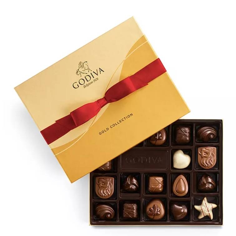 Godiva Chocolate Gift Box features 18 assorted chocolates in various shapes including hearts, crescents, and stars, displayed in an open box with a gold and red ribboned lid. The collection includes dark, milk, and white chocolates with various fillings.